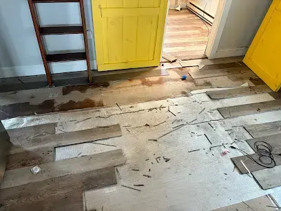 Wet floor with flooring boards pulled up beside a yellow door and a ladder