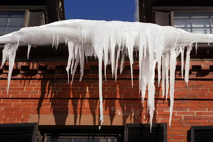 Heavy icicles and an ice ridge hanging from a snow-covered roof edge above a brick wall