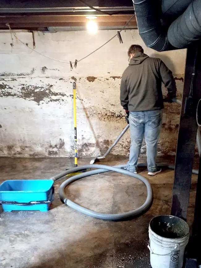 Technician running an extraction wand over a wet concrete basement floor beside a stained foundation wall