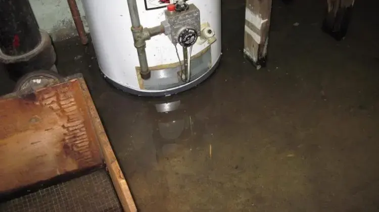 Water pooled on a dark basement floor around the base of a tank water heater